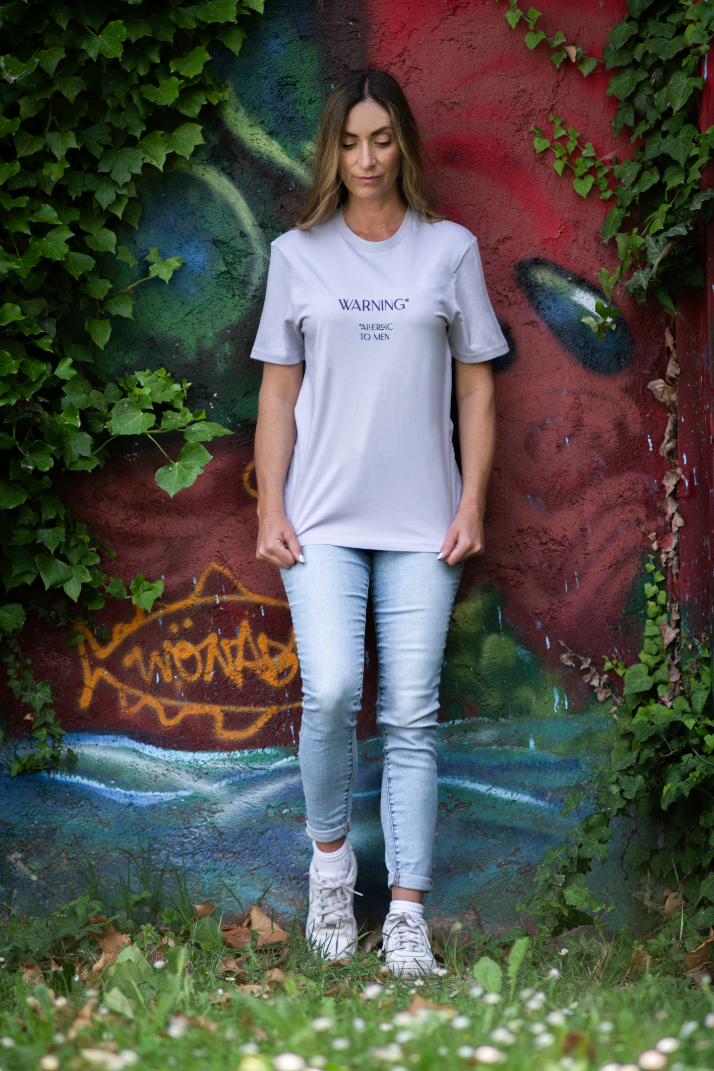Woman wearing lavender Allergic To Men t-shirt standing against colorful graffiti wall with ivy, blending urban and nature aesthetics