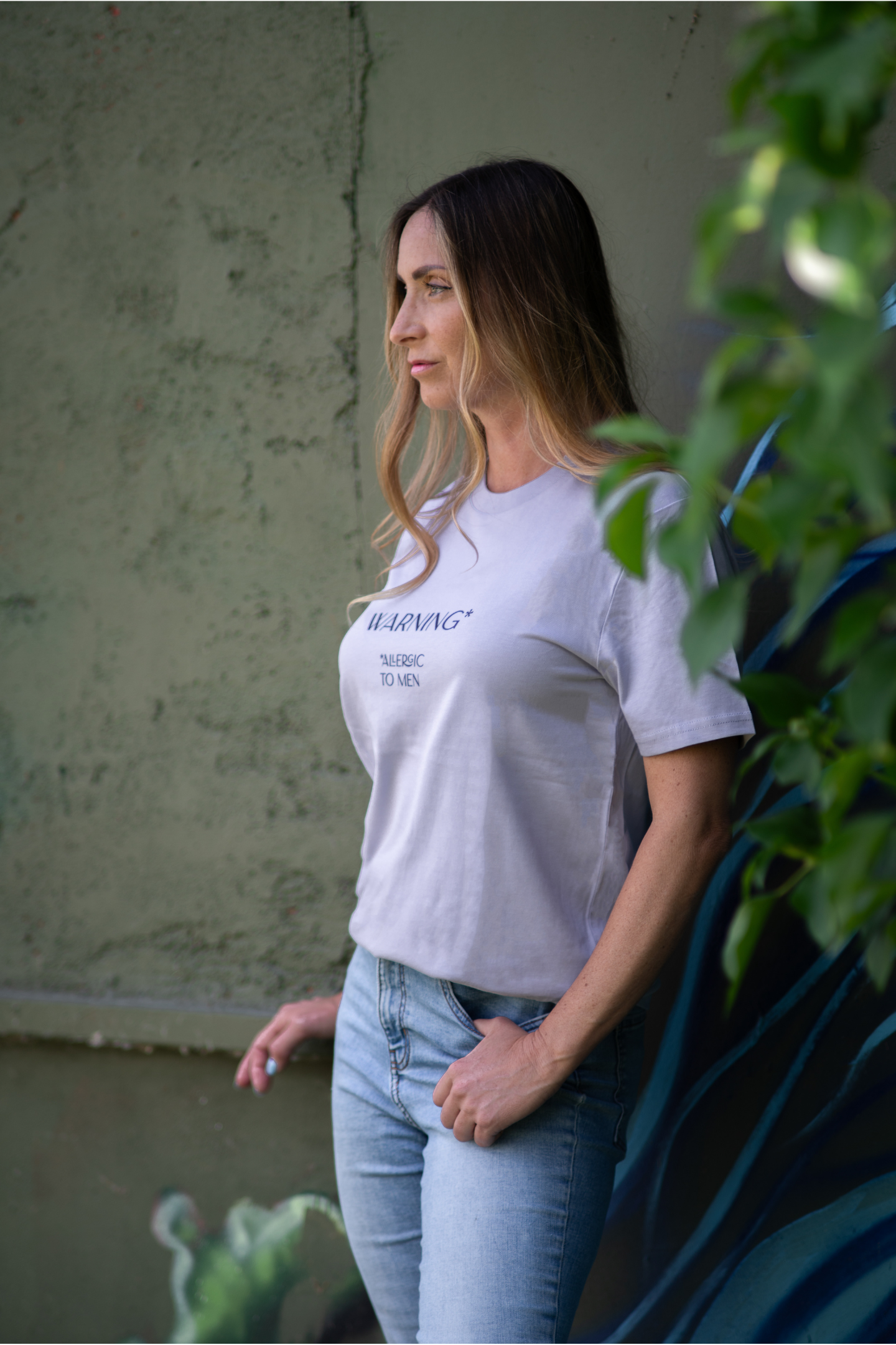 Woman in lavender Allergic To Men t-shirt posing side view near green wall with blue graffiti and leafy plant in foreground