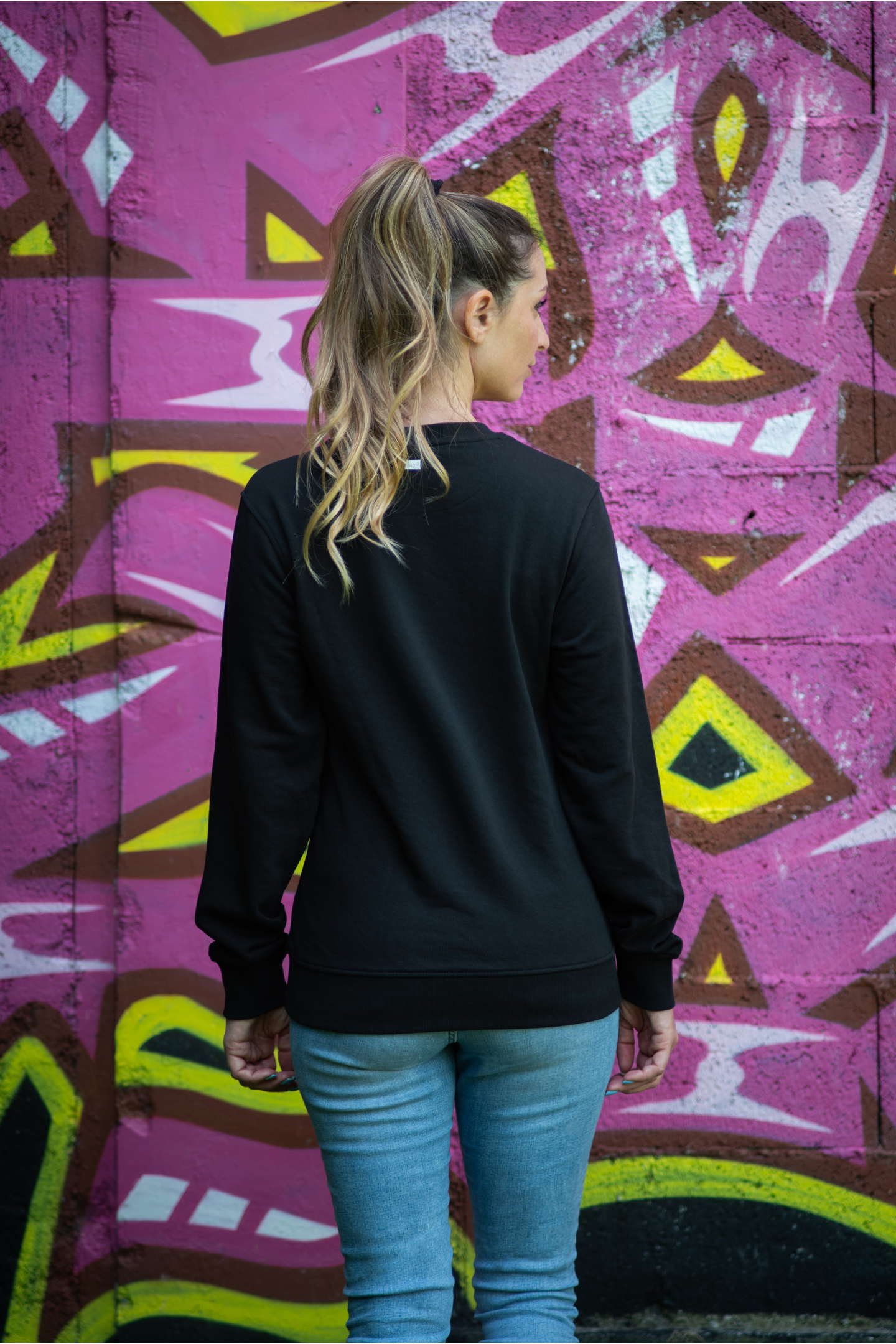 Back view of woman wearing black Danger unisex organic sweater with ponytail in front of bold pink graffiti wall
