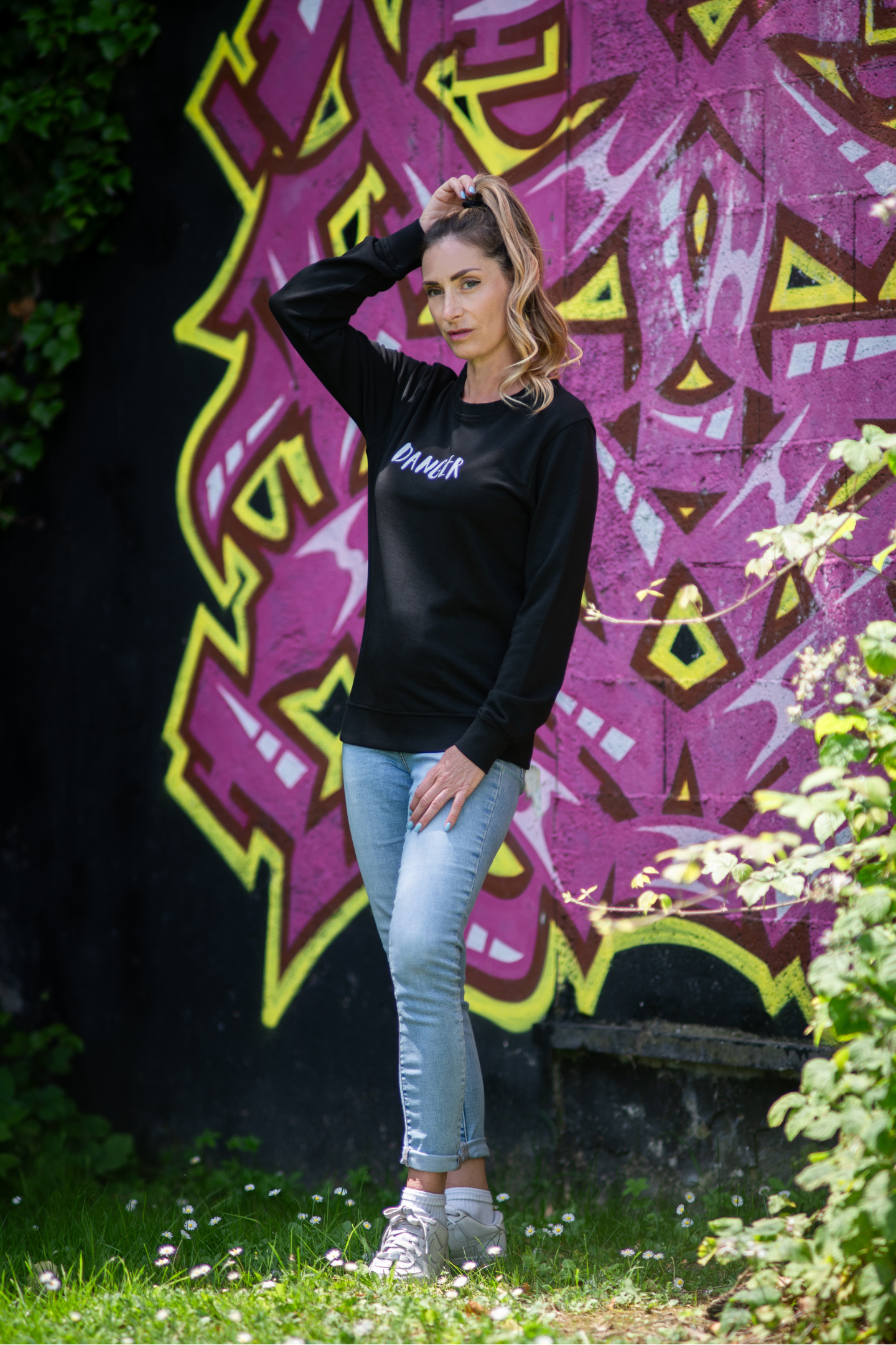 Woman posing outdoors in black Danger unisex organic sweater and light jeans, standing in front of colorful graffiti wall with hand in hair
