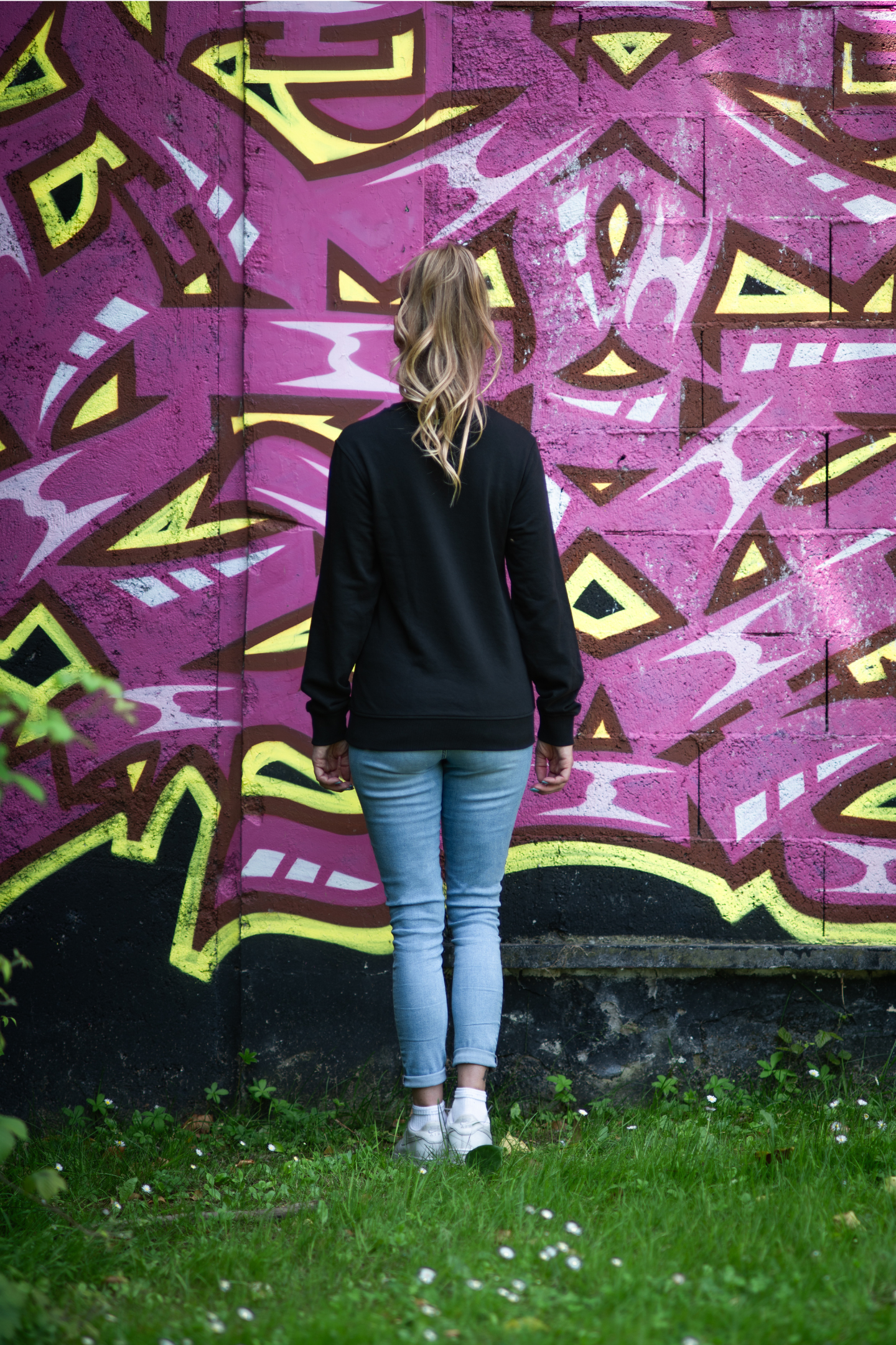 Woman standing in grass wearing black Danger unisex organic sweater with bold pink graffiti wall in background, full body back view