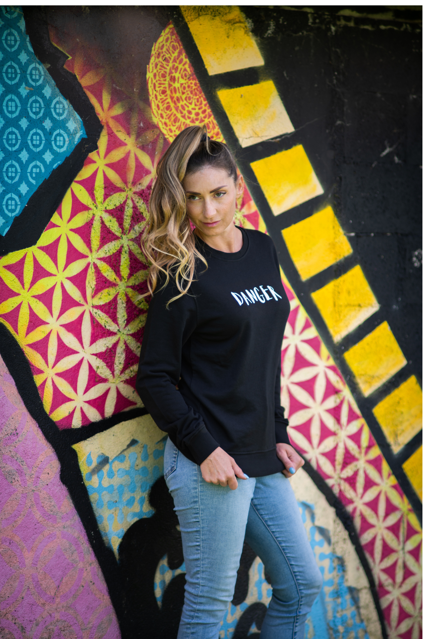 Woman wearing black Danger organic sweater posing against vibrant geometric graffiti wall