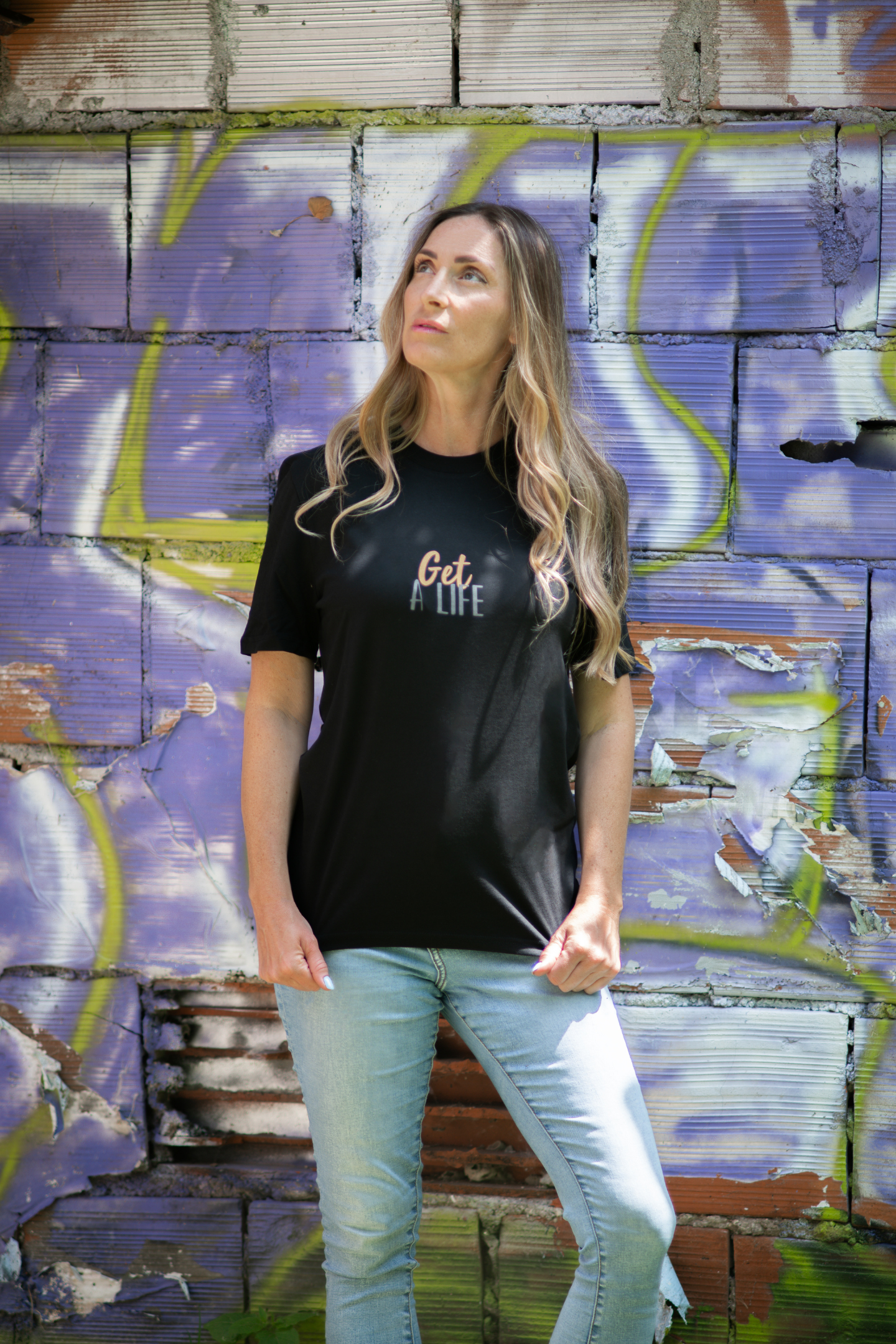 Woman wearing black Get A Life unisex organic tee in front of distressed purple graffiti brick wall, hands in pockets, looking upward