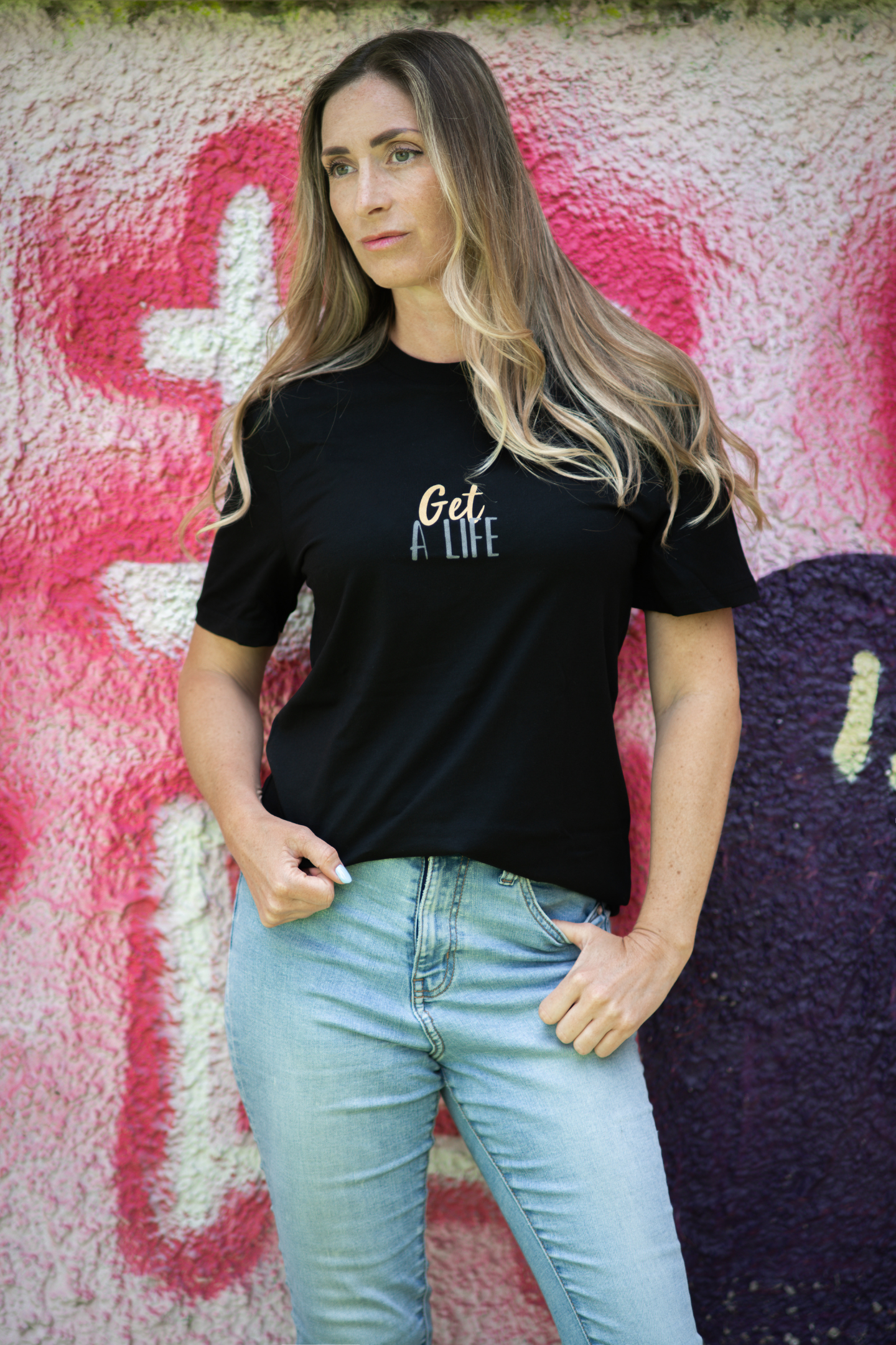 Woman in black Get A Life unisex tee facing forward, standing confidently against colorful graffiti wall