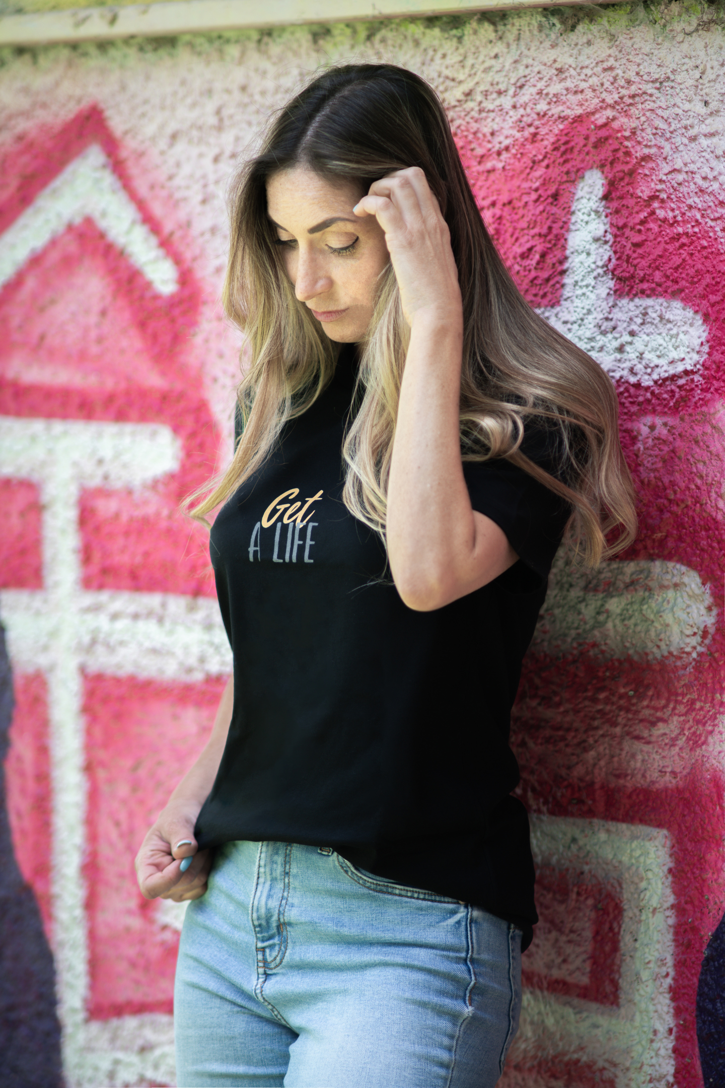Woman wearing black Get A Life unisex tee, looking down with hand in hair, standing by vibrant pink graffiti wall
