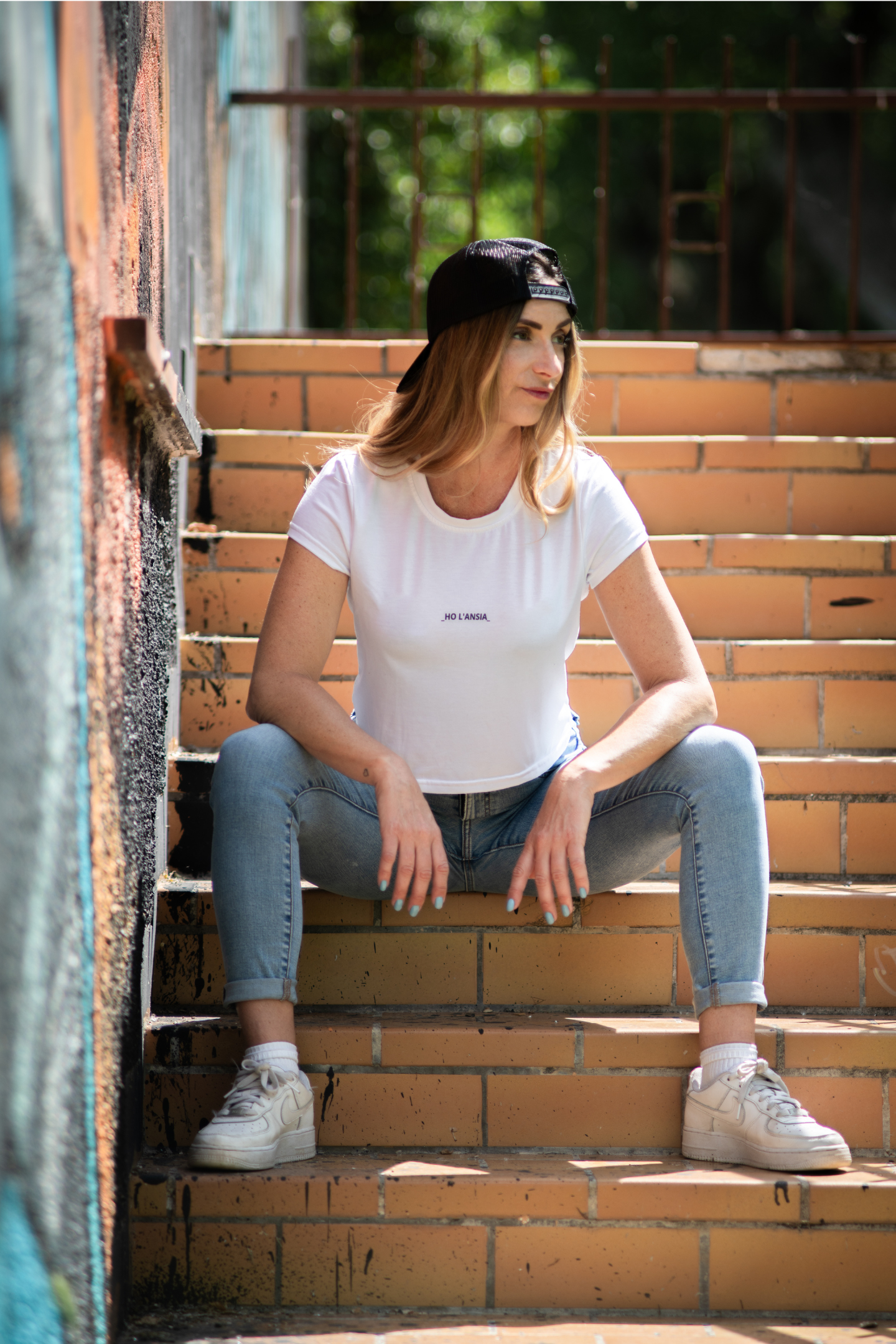 Model sitting on stairs wearing Ho L'Ansia Crop Tee with cap and white sneakers
