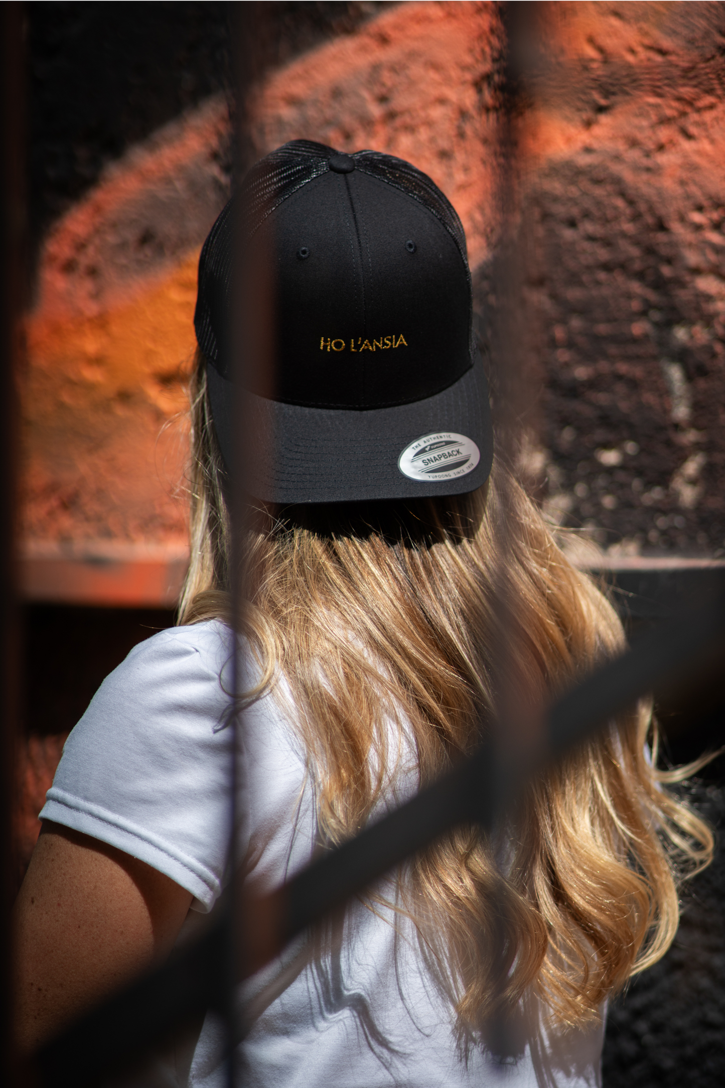 Blonde woman wearing Ho L'Ansia black mesh trucker cap with gold logo embroidery, photographed from behind
