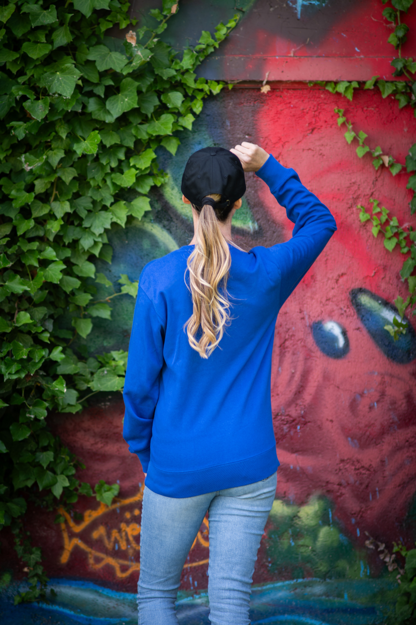 Back view of woman wearing blue Leave Me Alone sweatshirt and black cap in front of ivy-covered graffiti wall