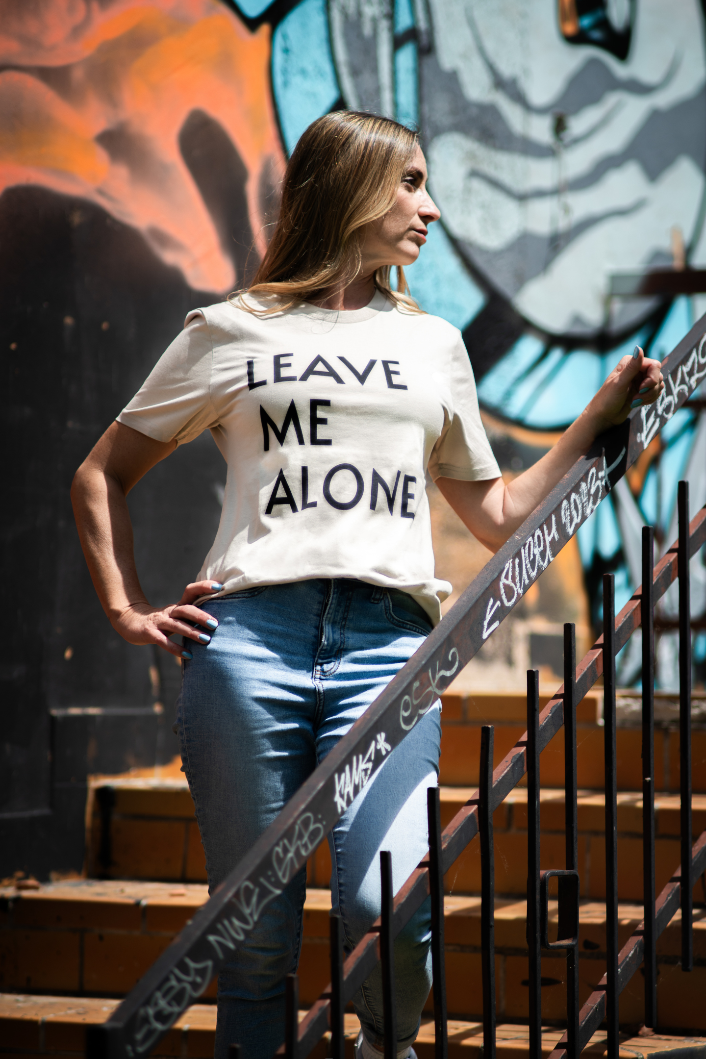 Woman wearing light Leave Me Alone tee standing on stairs with hand on graffiti-covered railing