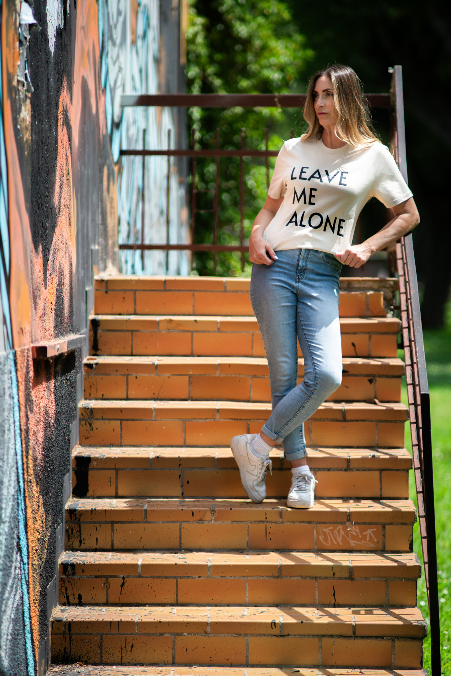 Woman in light Leave Me Alone unisex tee leaning on staircase railing with urban graffiti wall