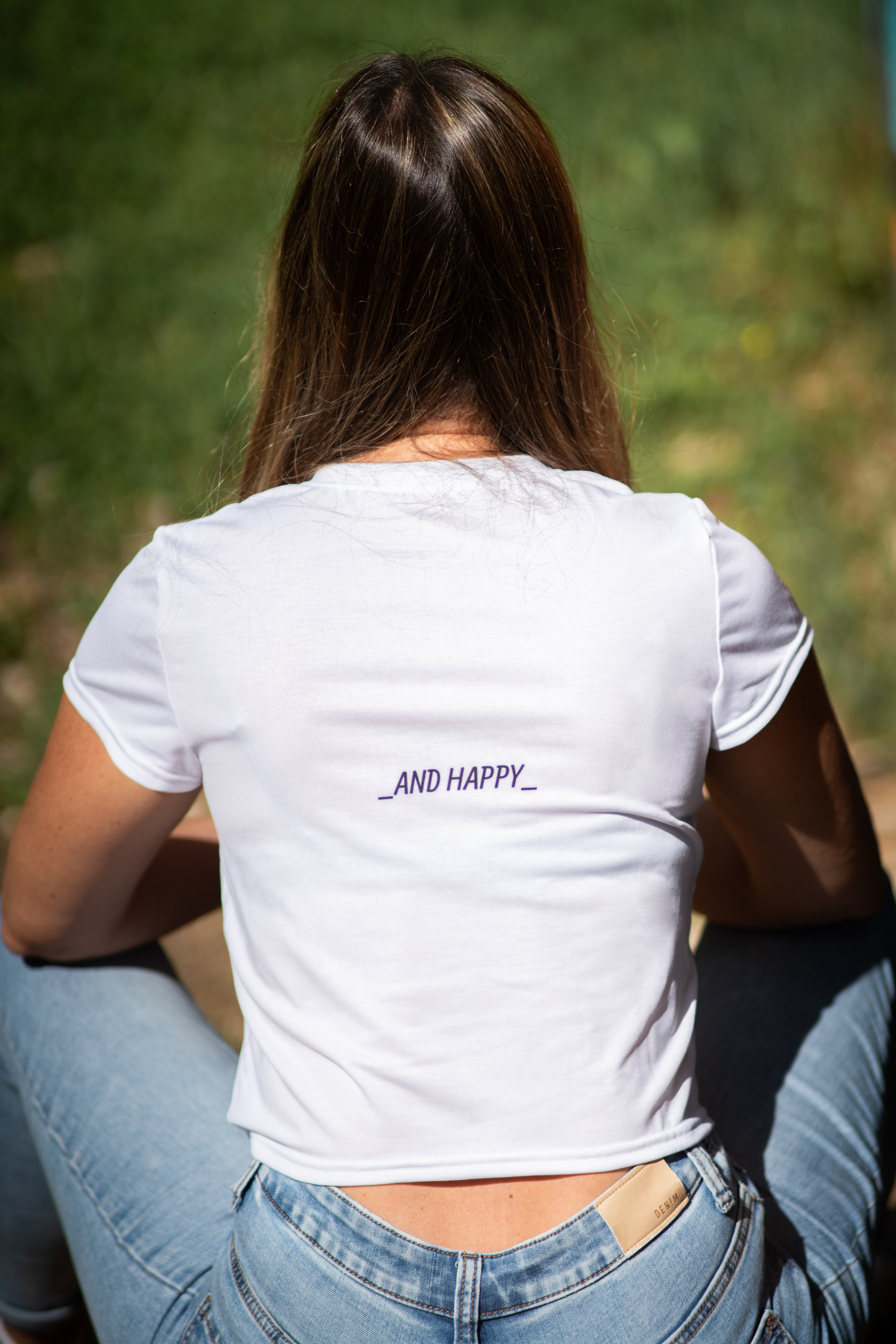 Close-up of back print on Silyp Single and Happy Crop Tee in white, showing AND HAPPY text, model sitting outdoors in sunlight