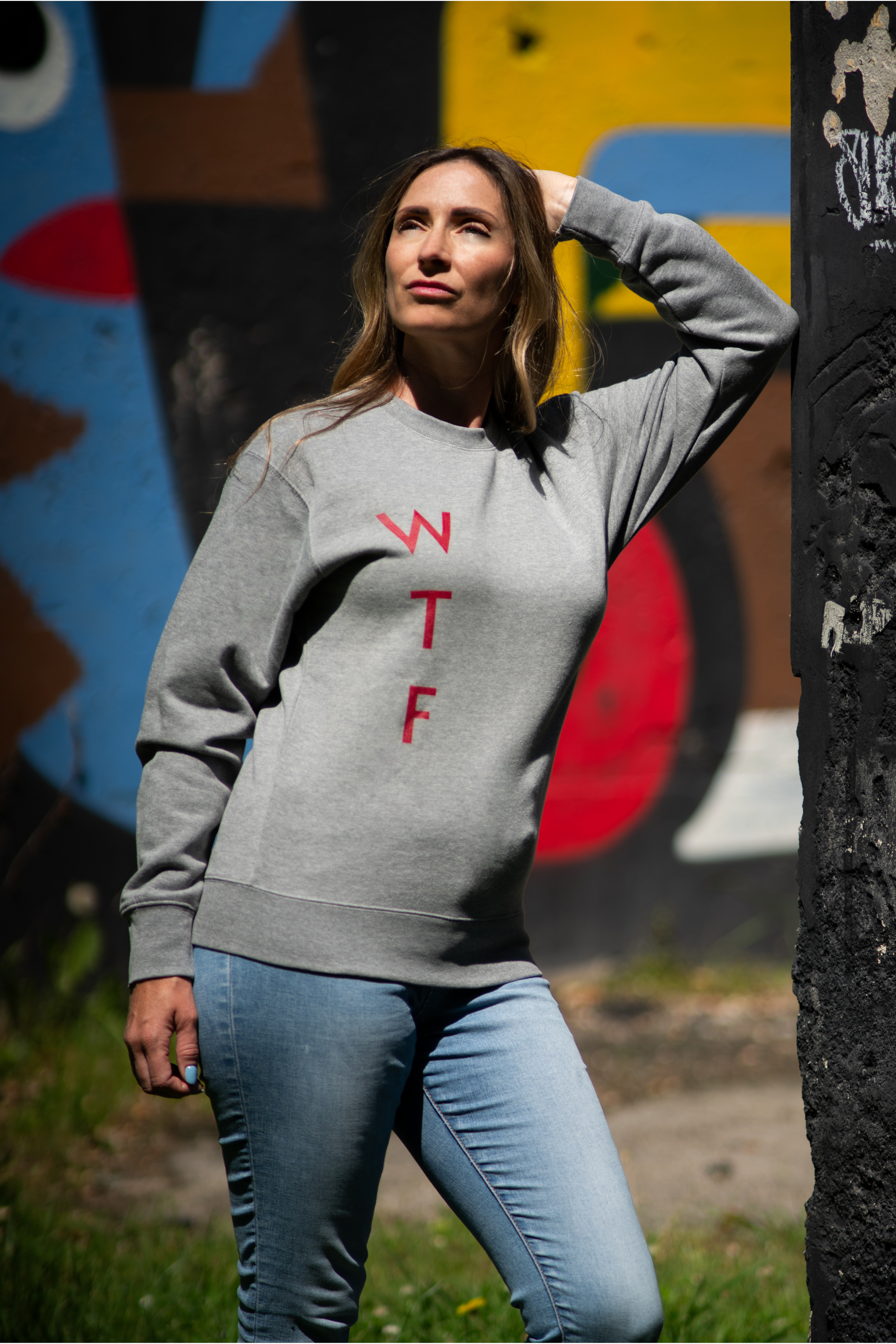 Woman posing in grey WTF sweatshirt with red print in front of colorful graffiti wall