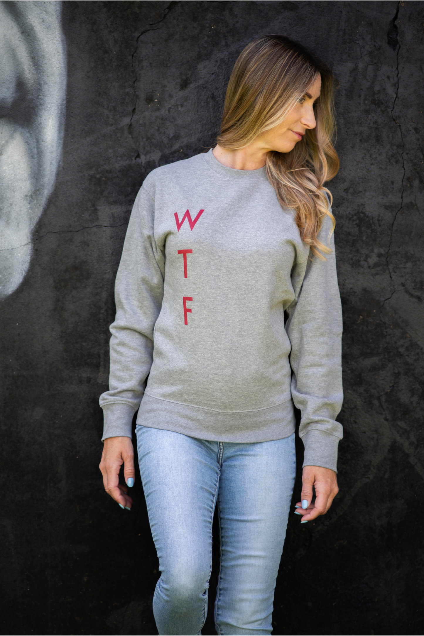 Woman wearing grey WTF unisex sweatshirt with red lettering, front view against dark graffiti wall