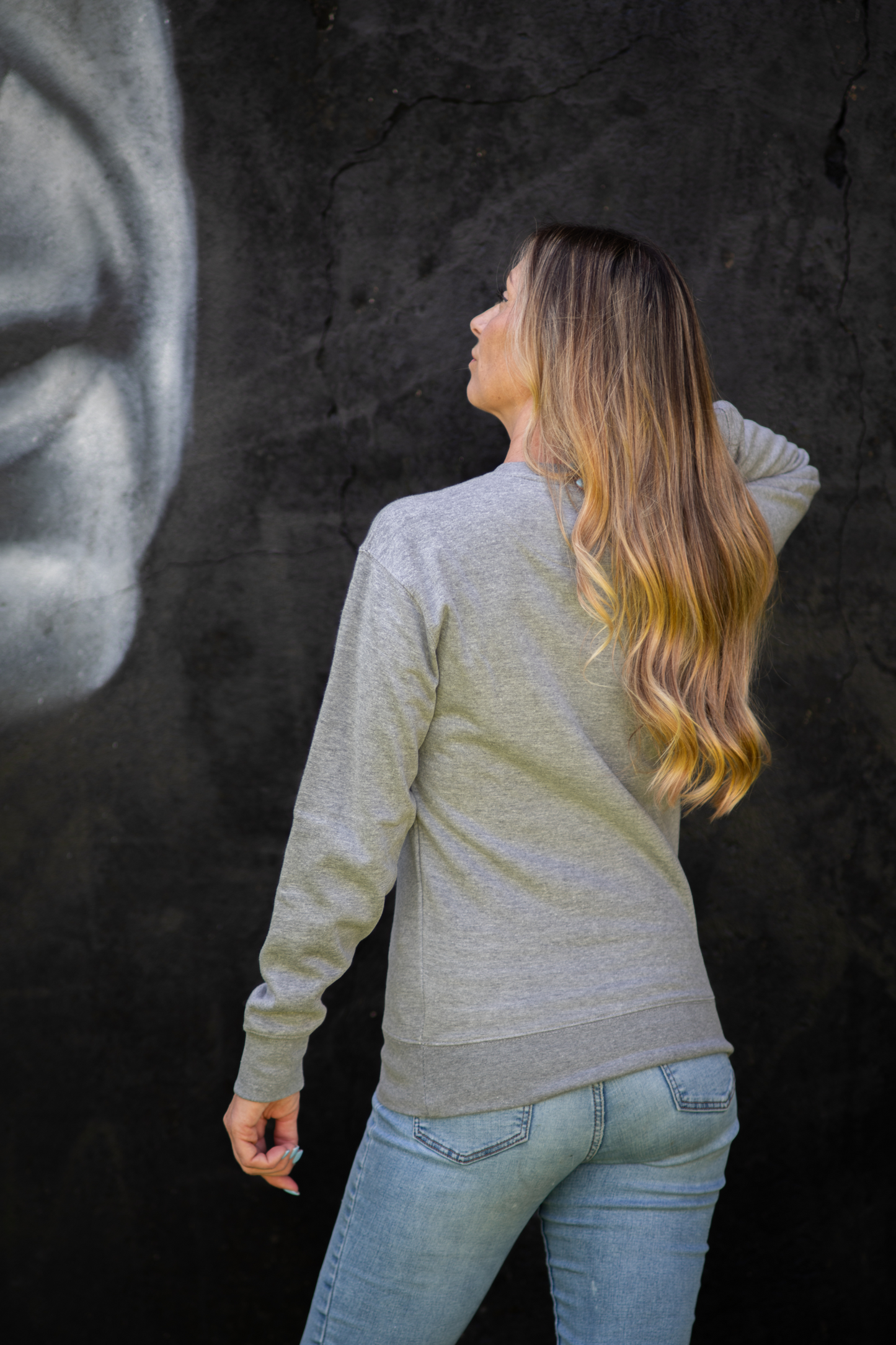 Back view of woman wearing heather white WTF unisex sweatshirt, posing in front of black graffiti wall