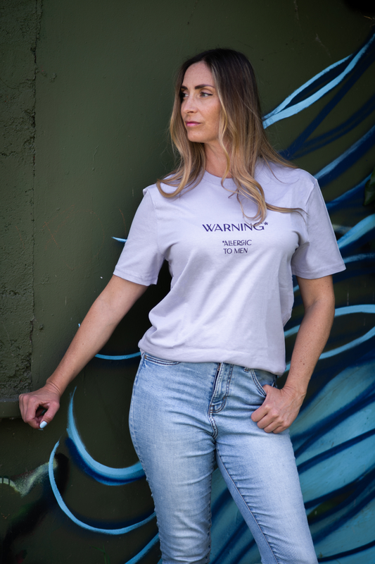 Woman wearing lavender Allergic To Men tee leaning against painted green wall with blue wave art, casual streetwear style