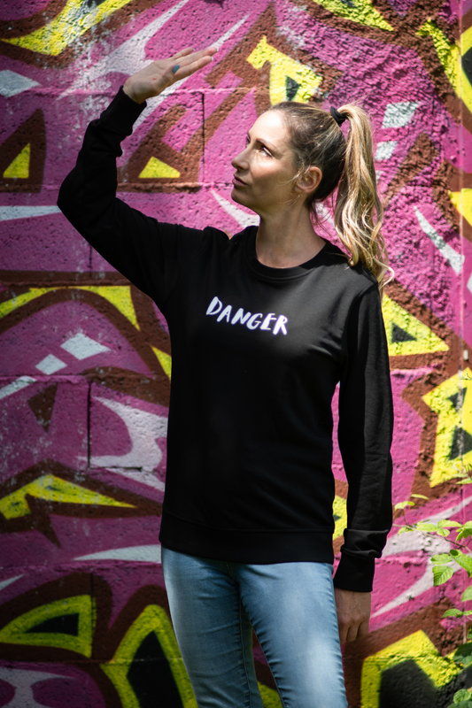 Model in black unisex Danger organic sweater shielding eyes from sunlight in front of vibrant graffiti wall