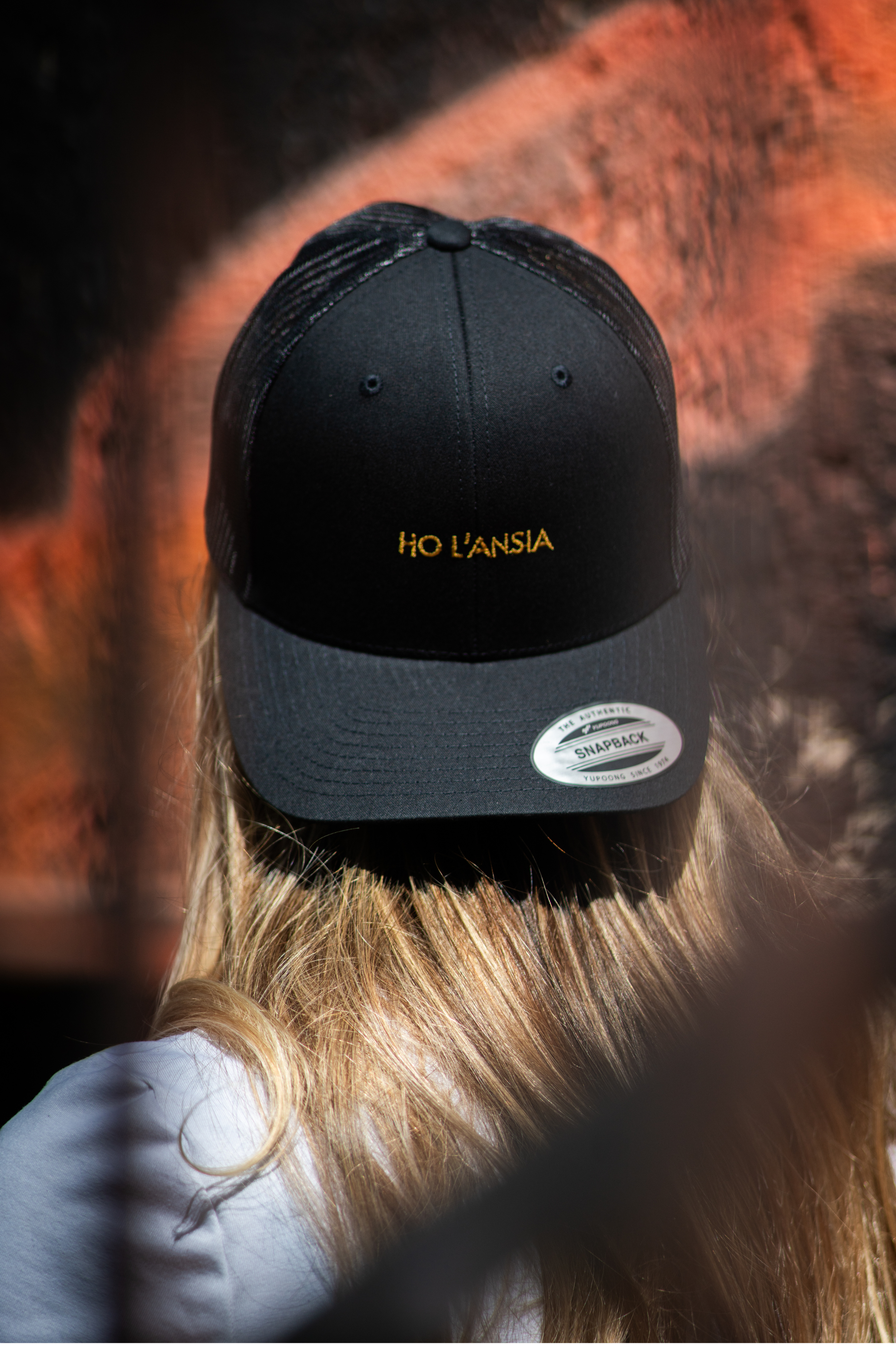 Close-up of Ho L'Ansia black trucker cap with gold embroidery, worn by blonde woman

