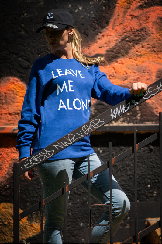 Model wearing blue Leave Me Alone unisex sweater and black cap on urban graffiti stairway