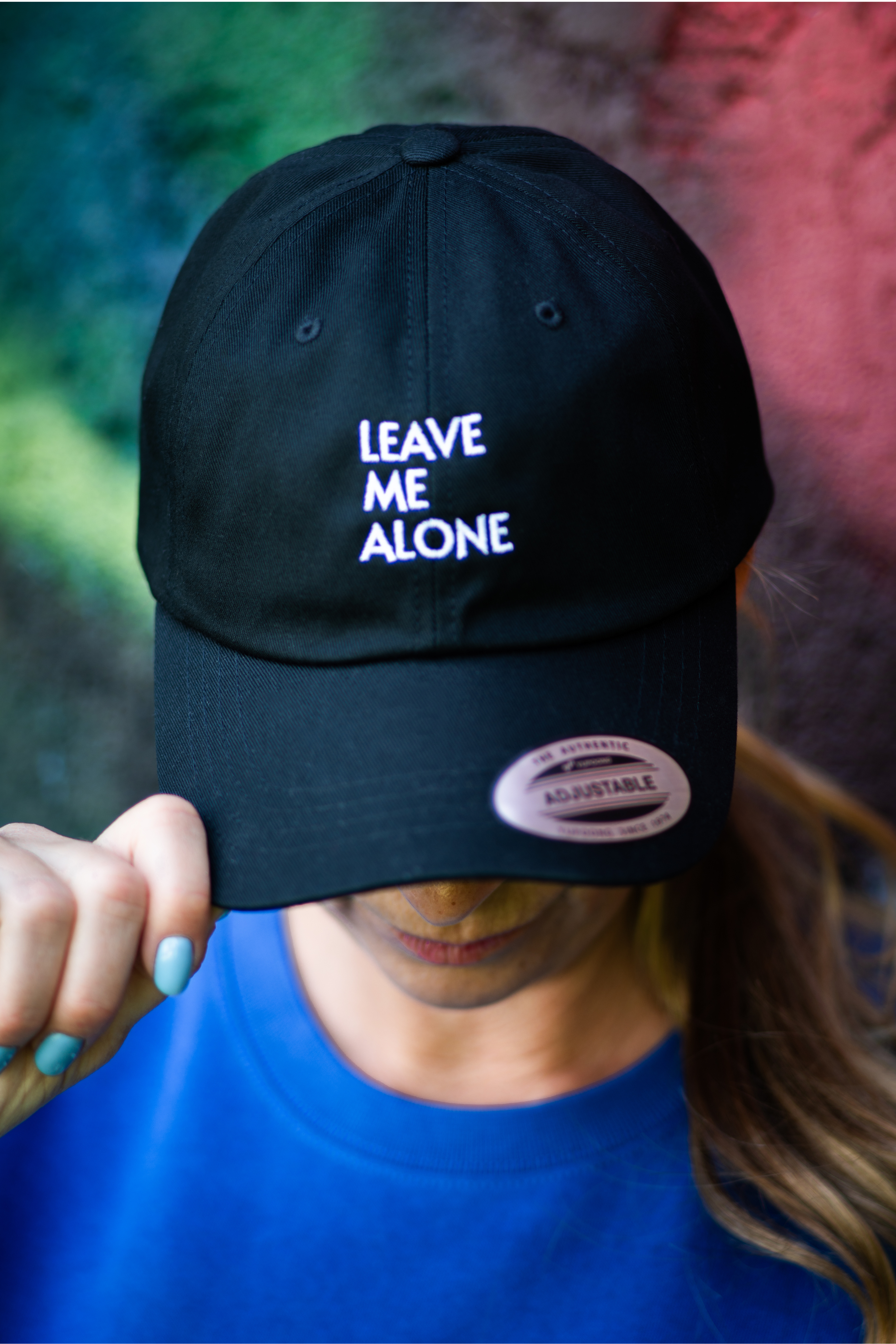 Close-up of black Leave Me Alone baseball cap with white embroidery, worn by woman in blue sweatshirt tilting the brim down