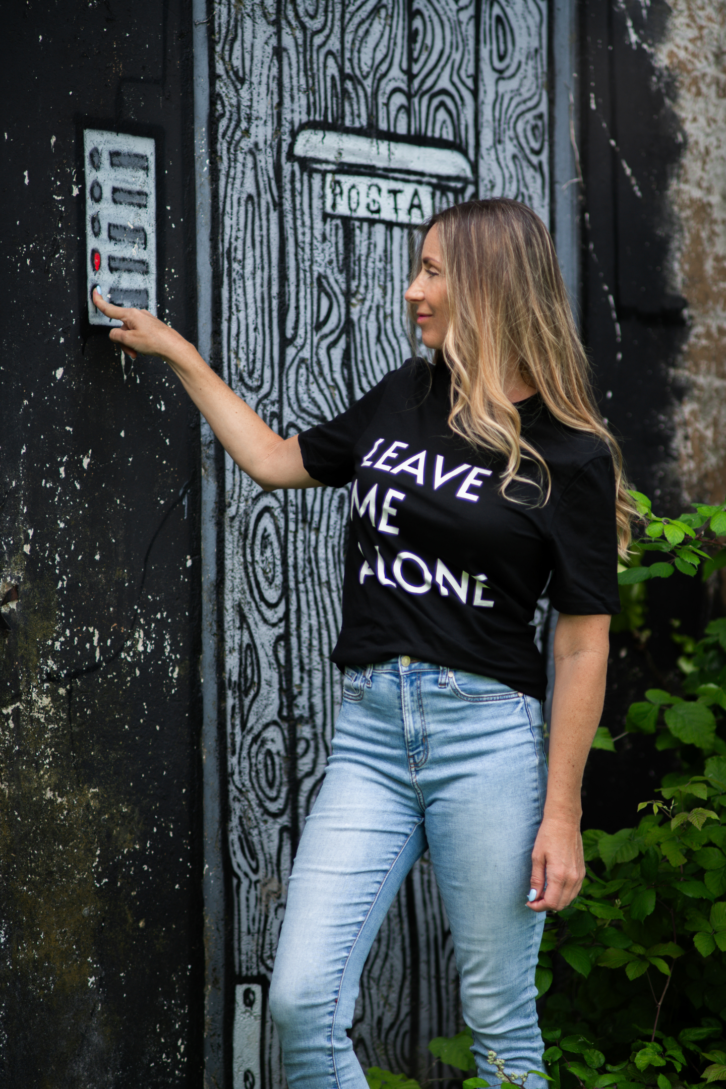 Woman wearing Leave Me Alone black tee pressing buzzer in front of painted door