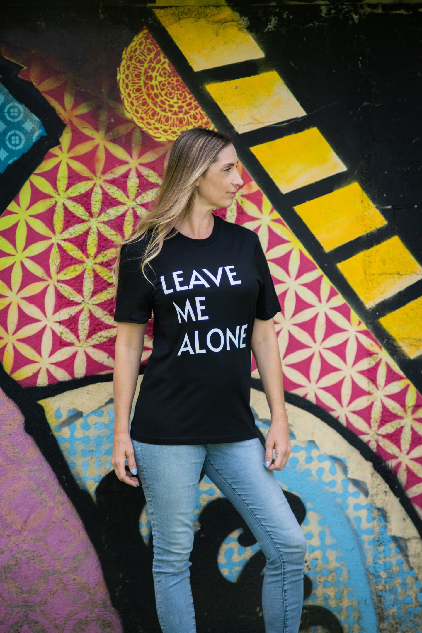 Woman wearing Leave Me Alone unisex tee, standing in front of vibrant graffiti wall with geometric patterns