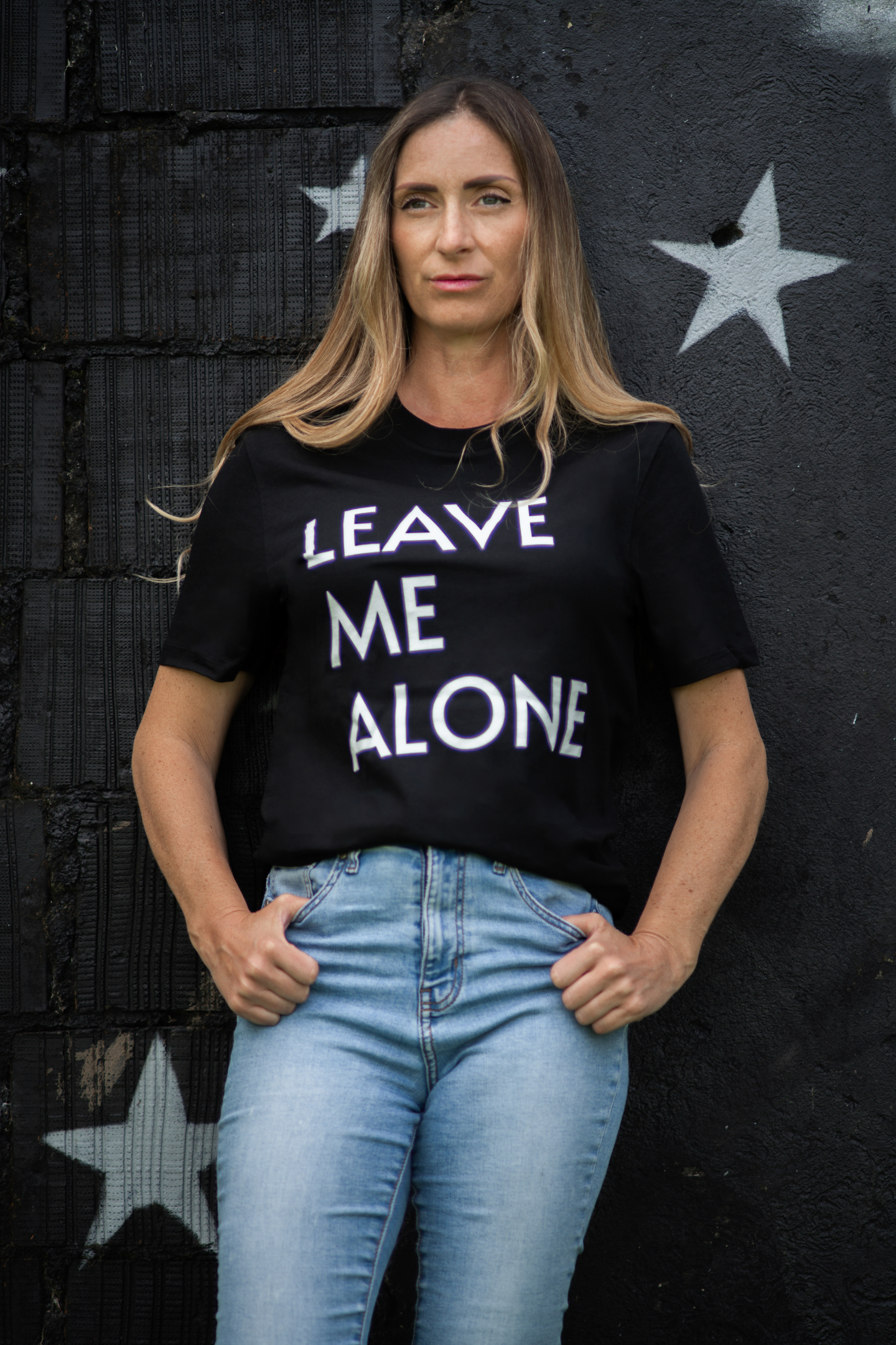 Woman wearing black Leave Me Alone unisex tee standing confidently against black wall with white stars