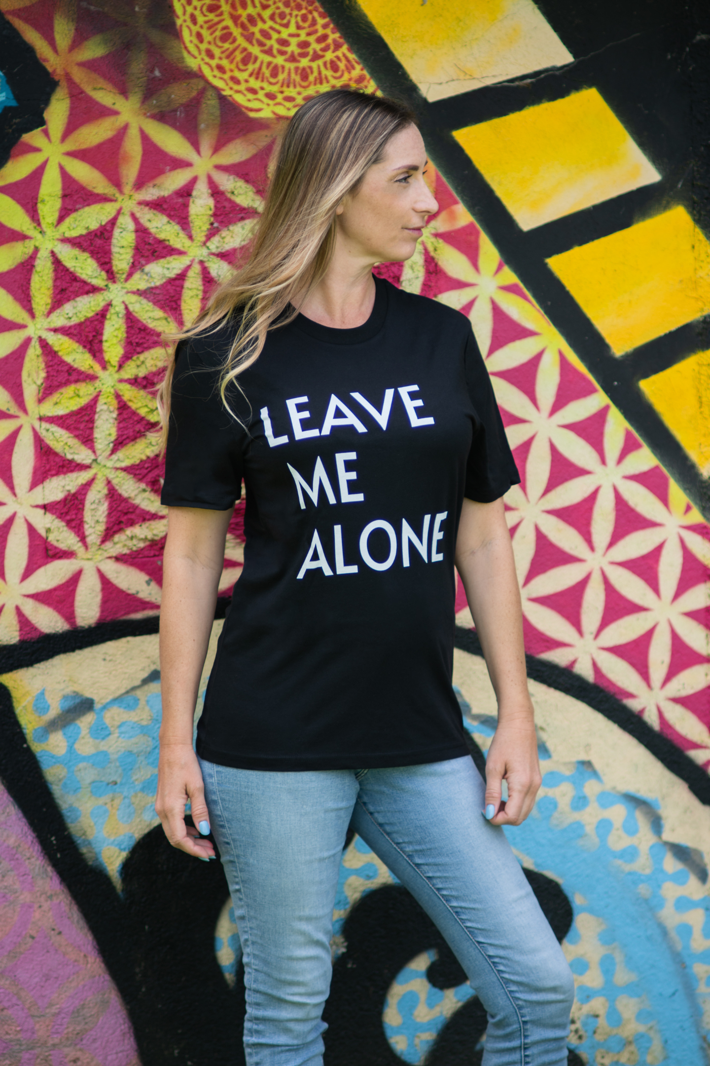 Woman in Leave Me Alone tee looking sideways, standing against vibrant geometric mural wall