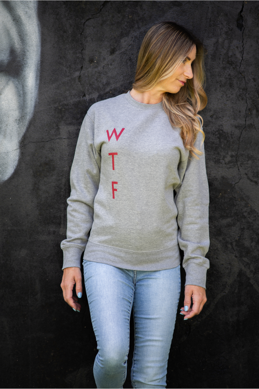 Woman wearing grey WTF unisex sweatshirt with red lettering, front view against dark graffiti wall