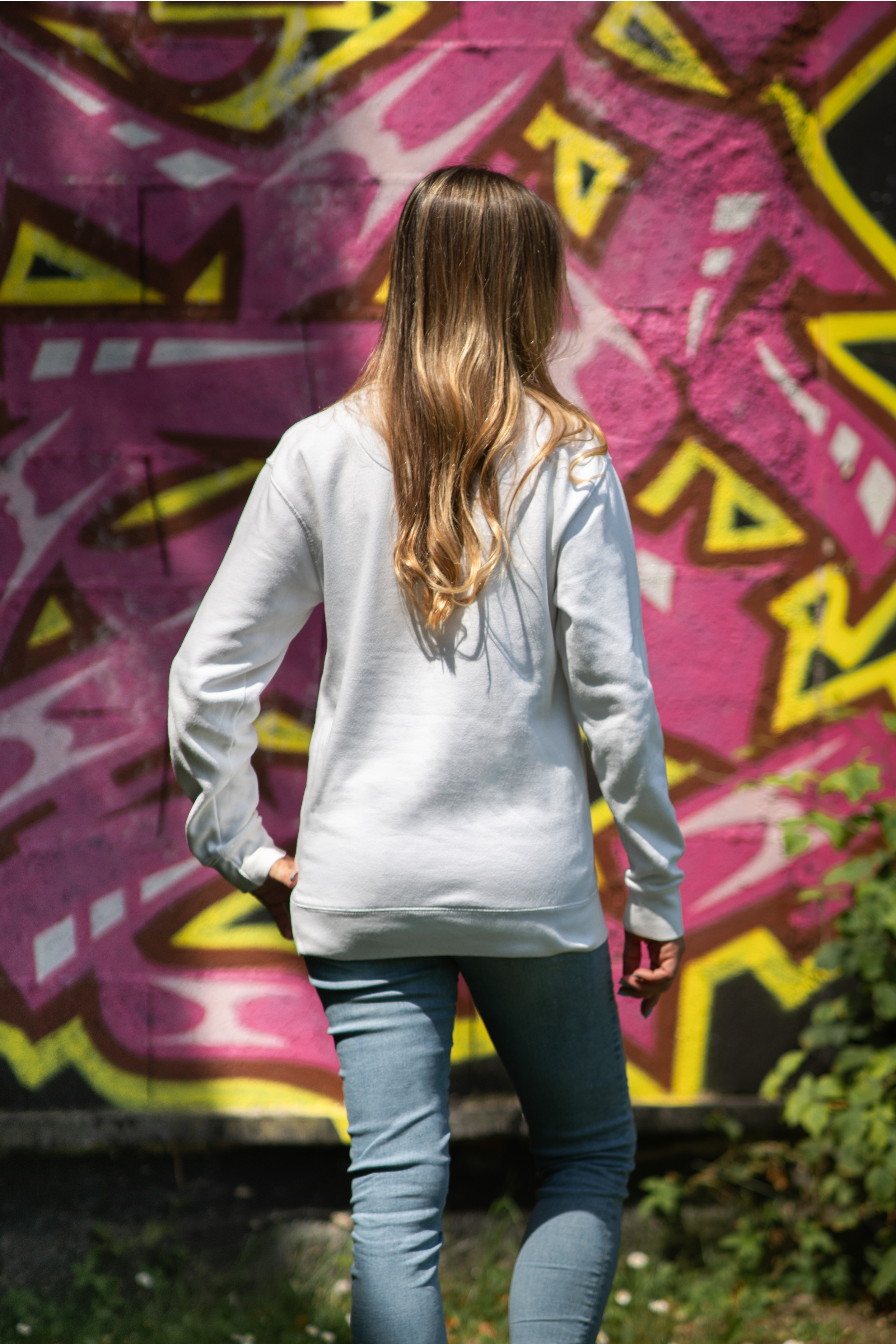 Back view of woman wearing white WTF unisex sweatshirt in front of pink graffiti wall