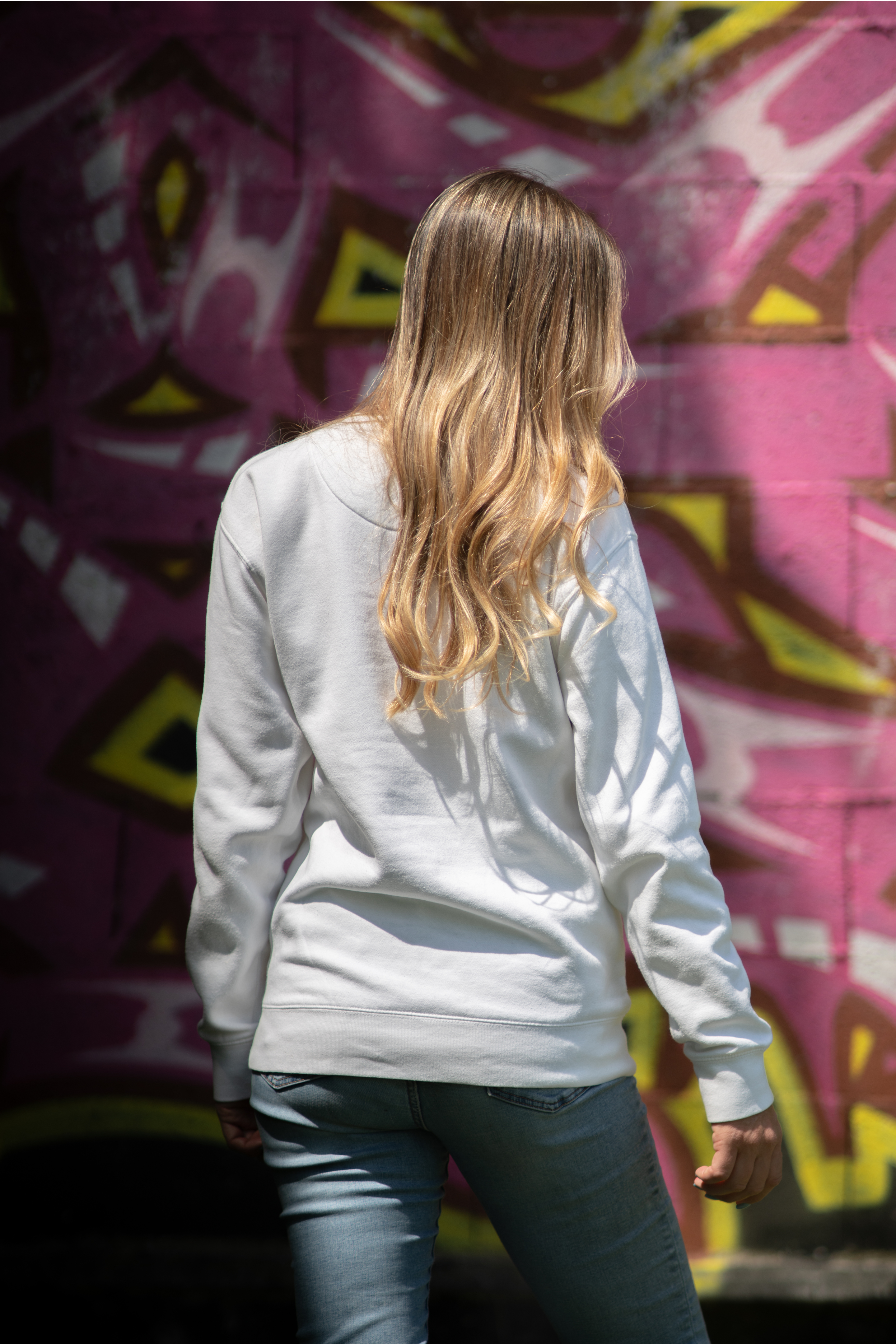 Back view of woman wearing white WTF unisex sweatshirt in front of vibrant pink graffiti wall