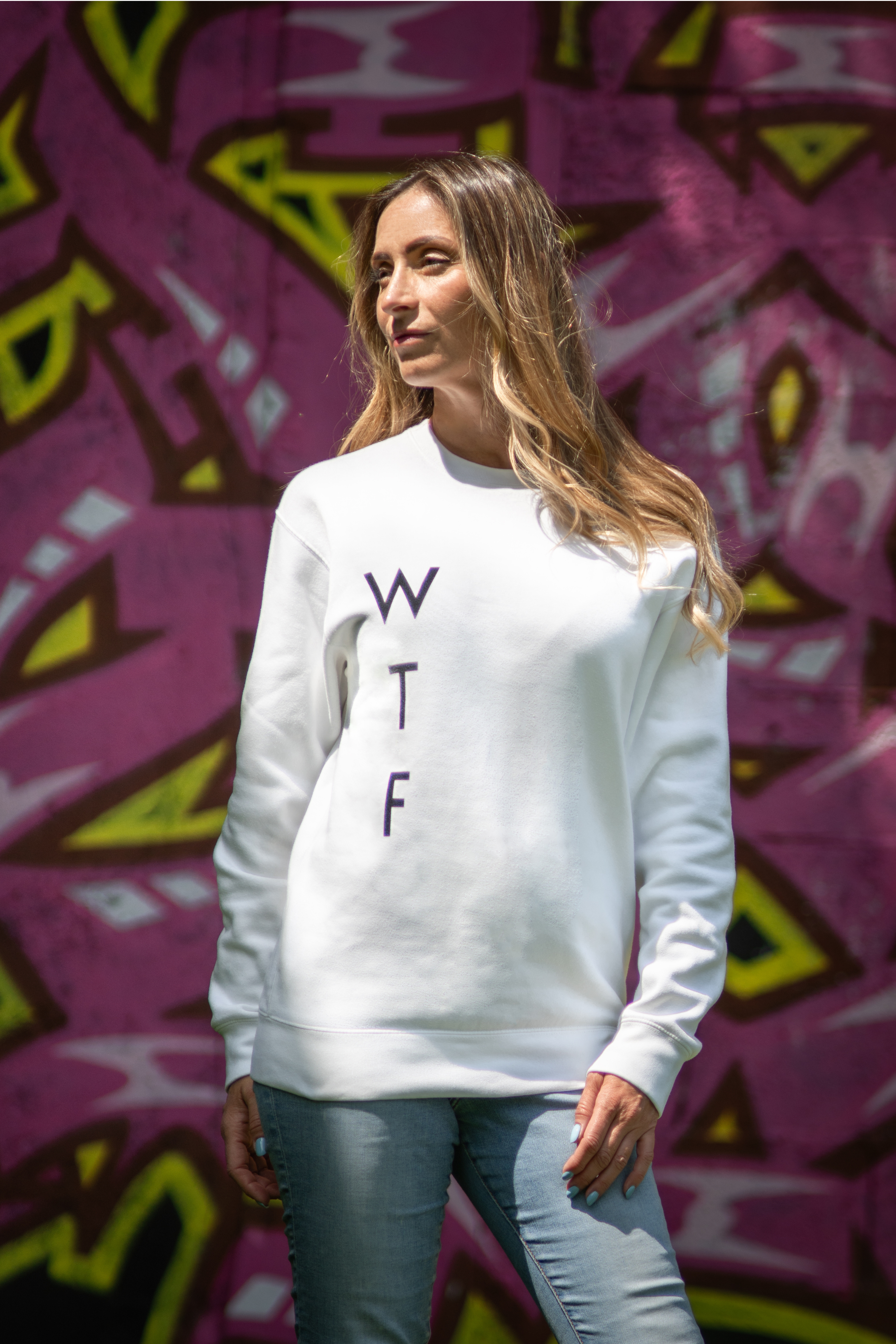 Woman wearing white WTF unisex sweatshirt in front of pink graffiti wall, front view